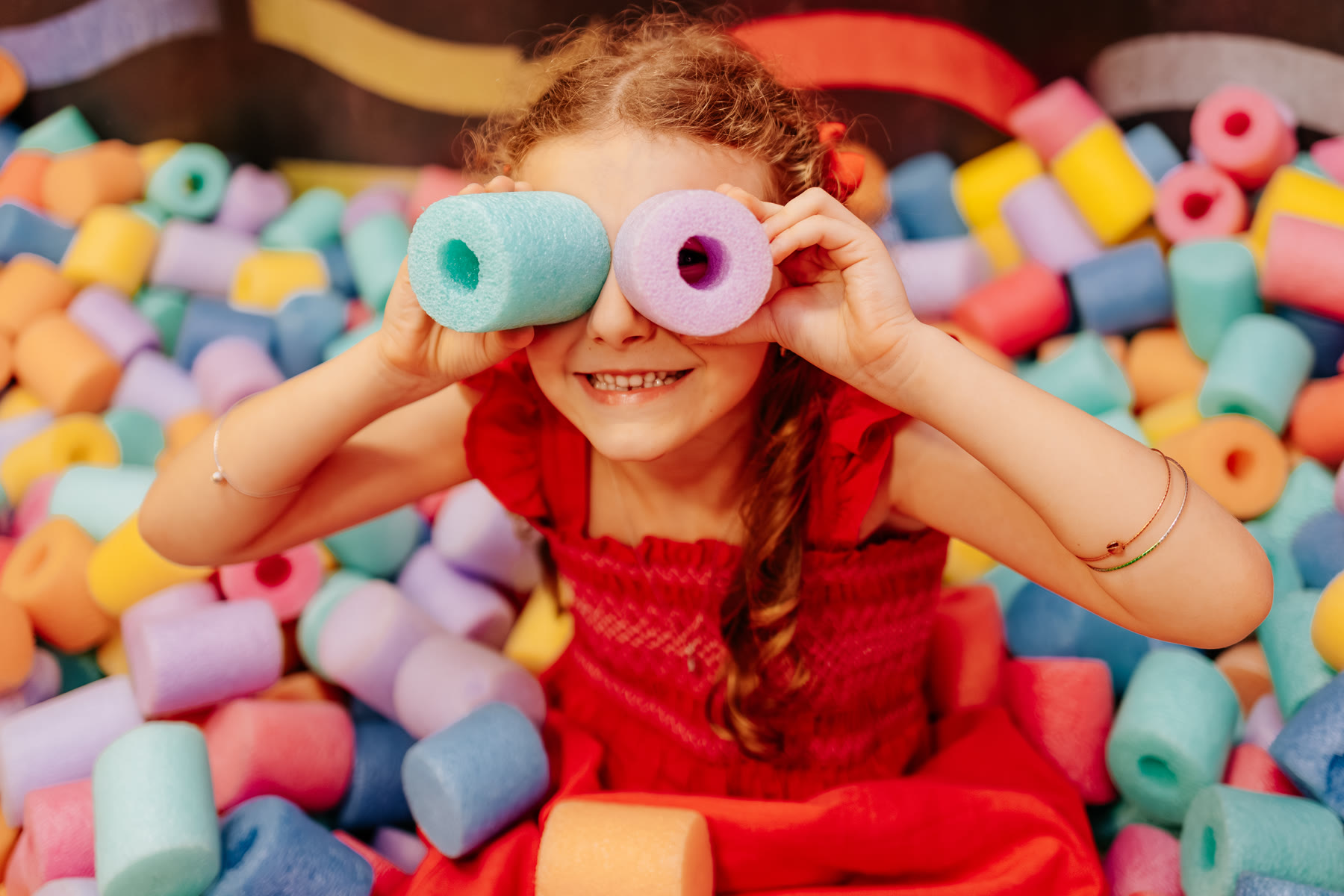 Menina sorrindo com espaguetes coloridos nos olhos, em piscina de espumas